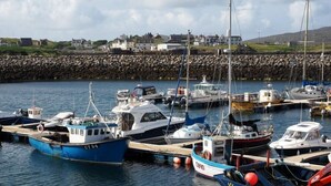 A harbor with boats docked, a stone breakwater, and houses in the background. - Tulloch House (South Uist)