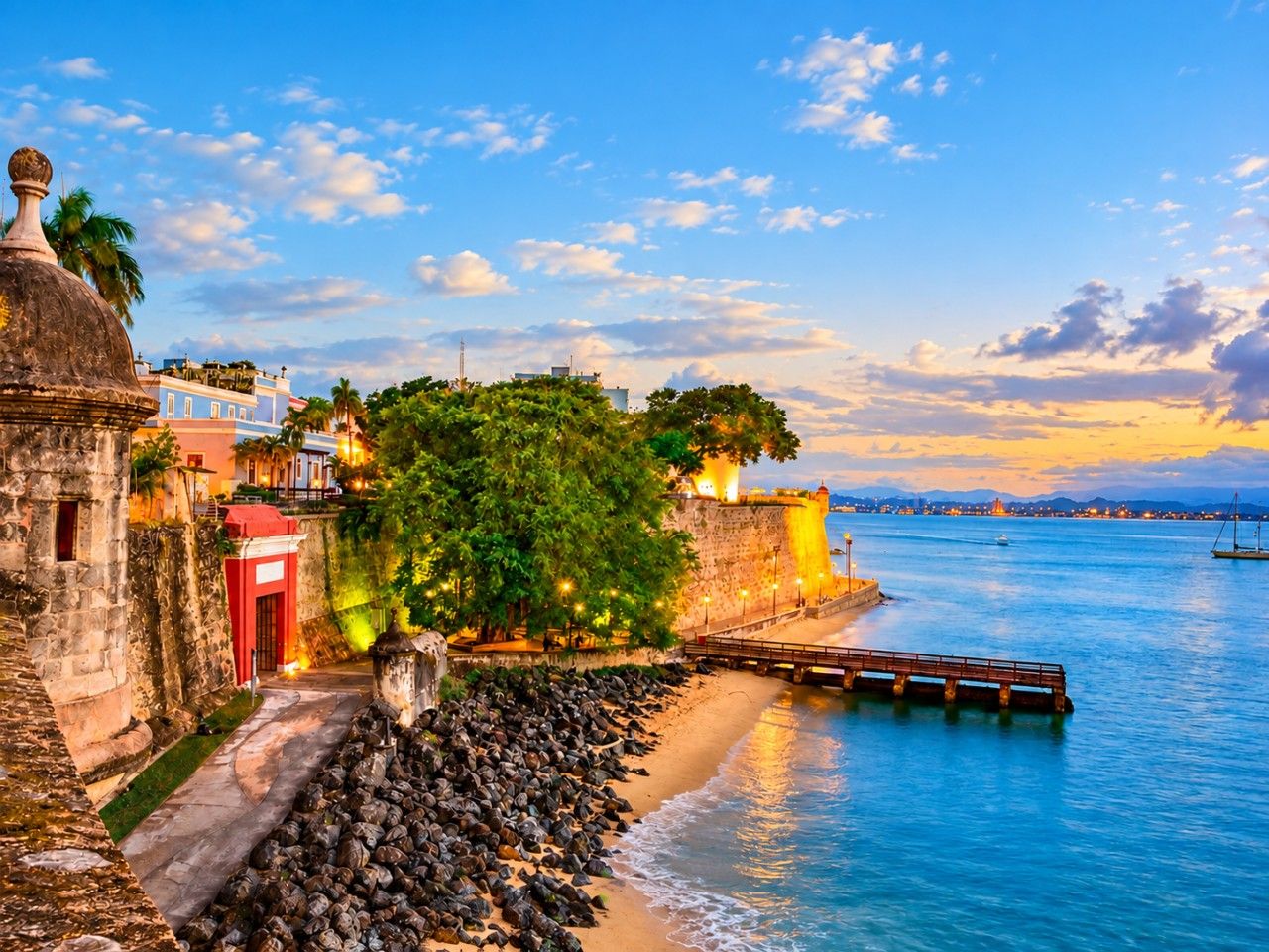 A coastal scene with a stone wall, a red door, and a bridge leading to a dock over calm waters.