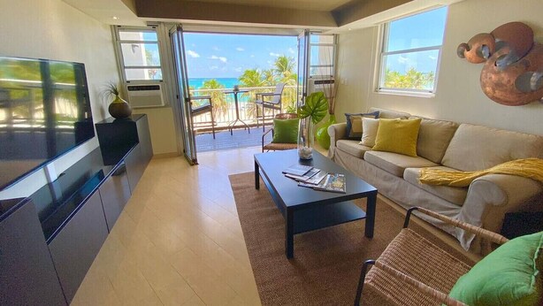 A modern living room with a large TV, a sofa, a coffee table, and a view of the ocean through large windows. - Beachfront 3-bedroom  Isla Verde  Walk 2 eat (Carolina)
