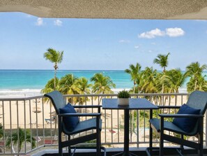 Un balcon avec deux chaises et une table, donnant sur une plage bordée de palmiers et sur l’océan.