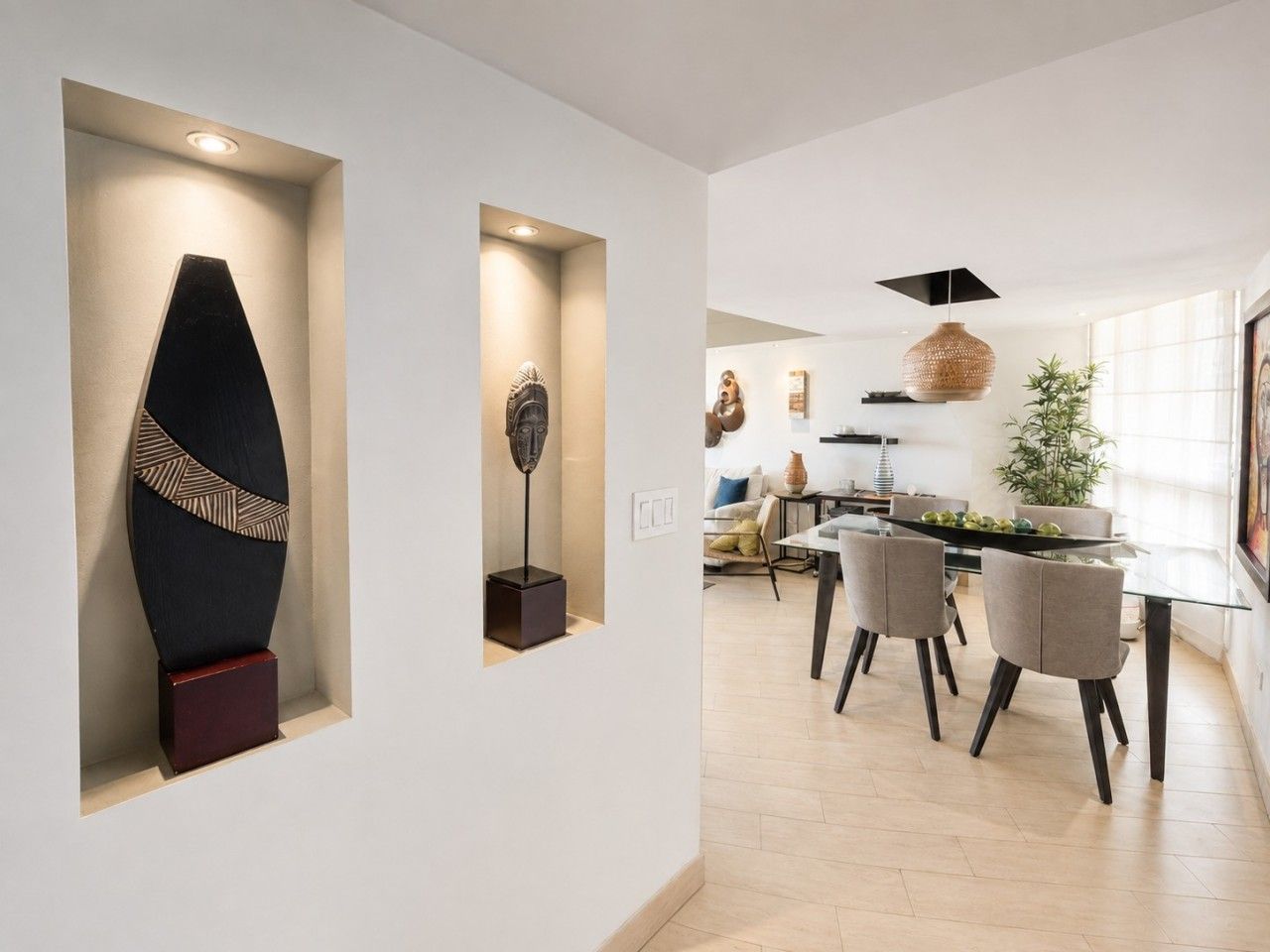 A modern interior with a dining area featuring a glass table and chairs, a kitchen with a hanging light fixture, and a wall with a geometric sculpture.