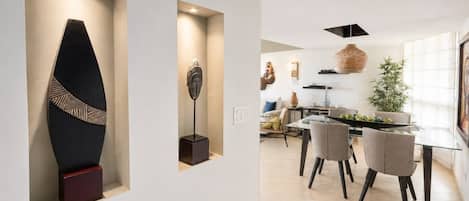 A modern interior with a dining area featuring a glass table and chairs, a kitchen with a hanging light fixture, and a wall with a geometric sculpture.