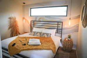 Un dormitorio moderno con una cama grande, un mural decorativo, una ventana y una pequeña mesa redonda con una planta.
