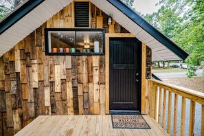 Una cabaña de madera rústica con una puerta negra, una ventana con tazas de colores y un felpudo en la terraza.