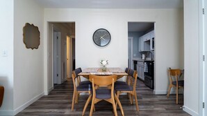 A dining area with a wooden table and chairs, a clock on the wall, and a kitchen in the background. - Madrona's Cottage B- Your Central Hub to Charleston! 5 Min From Airport (North Charleston)