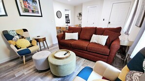 A living room with a red sofa, a multi-colored armchair, and a round ottoman. There is a dining area in the background with a white door and a clock on the wall. - Madrona's Cottage B- Your Central Hub to Charleston! 5 Min From Airport (North Charleston)