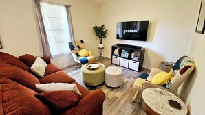 A living room with a large brown sofa, a yellow armchair, a small round ottoman, a marble coffee table, and a television on the wall. - Madrona's Cottage B- Your Central Hub to Charleston! 5 Min From Airport (North Charleston)