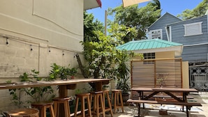 Ein Patio im Freien mit Holztischen und -hocker, umgeben von Grünpflanzen und einem Gebäude mit grauem Mauerwerk.
