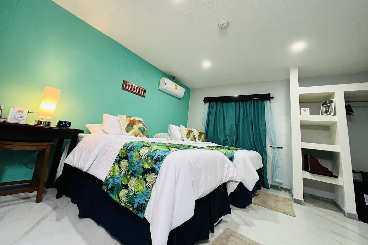 A hotel room with a bed, a desk with a lamp, a clock, and a shelf with items.