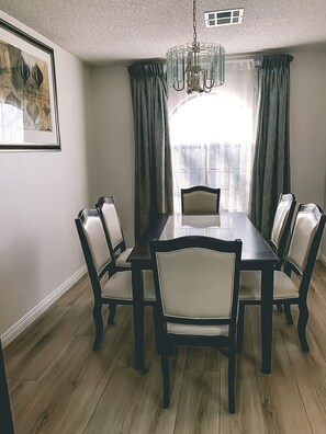 Enjoy meals in this dining room with a large table and six chairs, a chandelier, and a window with curtains.