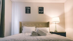 Un lit bien fait, avec du linge de lit blanc et des oreillers, un banc recouvert d’un tissu vert placé au pied du lit, un tableau illustrant différentes plantes aromatiques accroché au mur, et une table de chevet avec une lampe.