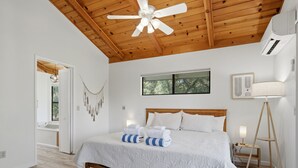 Camera da letto con soffitto in legno, un letto con biancheria bianca, una lampada da terra e una finestra vista alberi.