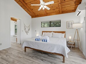 A bedroom with a wooden ceiling, a bed with white bedding, a standing lamp, and a window with a view of trees.