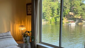A bedroom with a large window overlooking a lake, a bed with a textured blanket, a bedside lamp, and a potted plant. - Adorable Lake Getaway (Hot Springs)