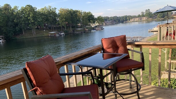 A deck with two chairs and a table overlooking a river with boats and buildings in the background. - Adorable Lake Getaway (Hot Springs)