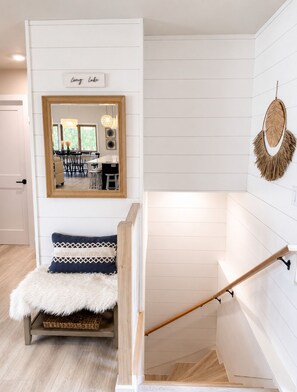 A staircase with a wooden handrail, a white wall with horizontal paneling, a mirror reflecting a dining area, and a sign above the mirror.