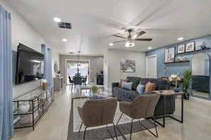 A modern living room with a sofa, coffee table, and chairs. There is a TV mounted on the wall, a ceiling fan, and a dining area in the background.