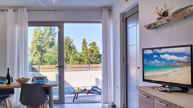 Ein modernes Wohnzimmer mit großem Fernseher, Esstisch und Blick auf einen Strand durch eine Schiebetür.