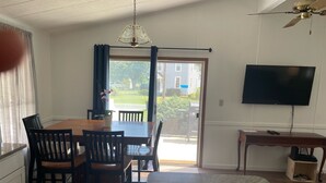Ein Essbereich mit Holztisch und Stühlen, einem Deckenventilator, einem an der Wand montierten Fernseher und Blick ins Freie durch eine Glastür.