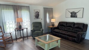 Ein Wohnzimmer mit einem grünen Sessel, einem braunen Ledersofa, einem Glastisch und zwei Lampen auf Beistelltischen.