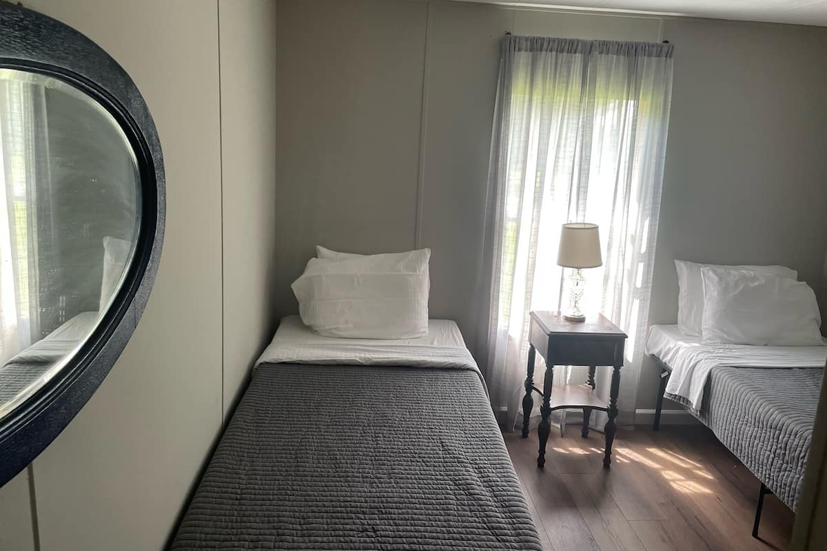 A room with two single beds, a small table with a lamp, a round mirror, and a window with sheer curtains.