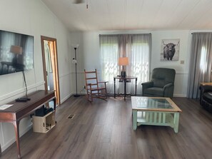 A living room with a wooden floor, a television mounted on the wall, a rocking chair, a green armchair, a glass coffee table, and a lamp on a side table. - West End House (Wachapreague)