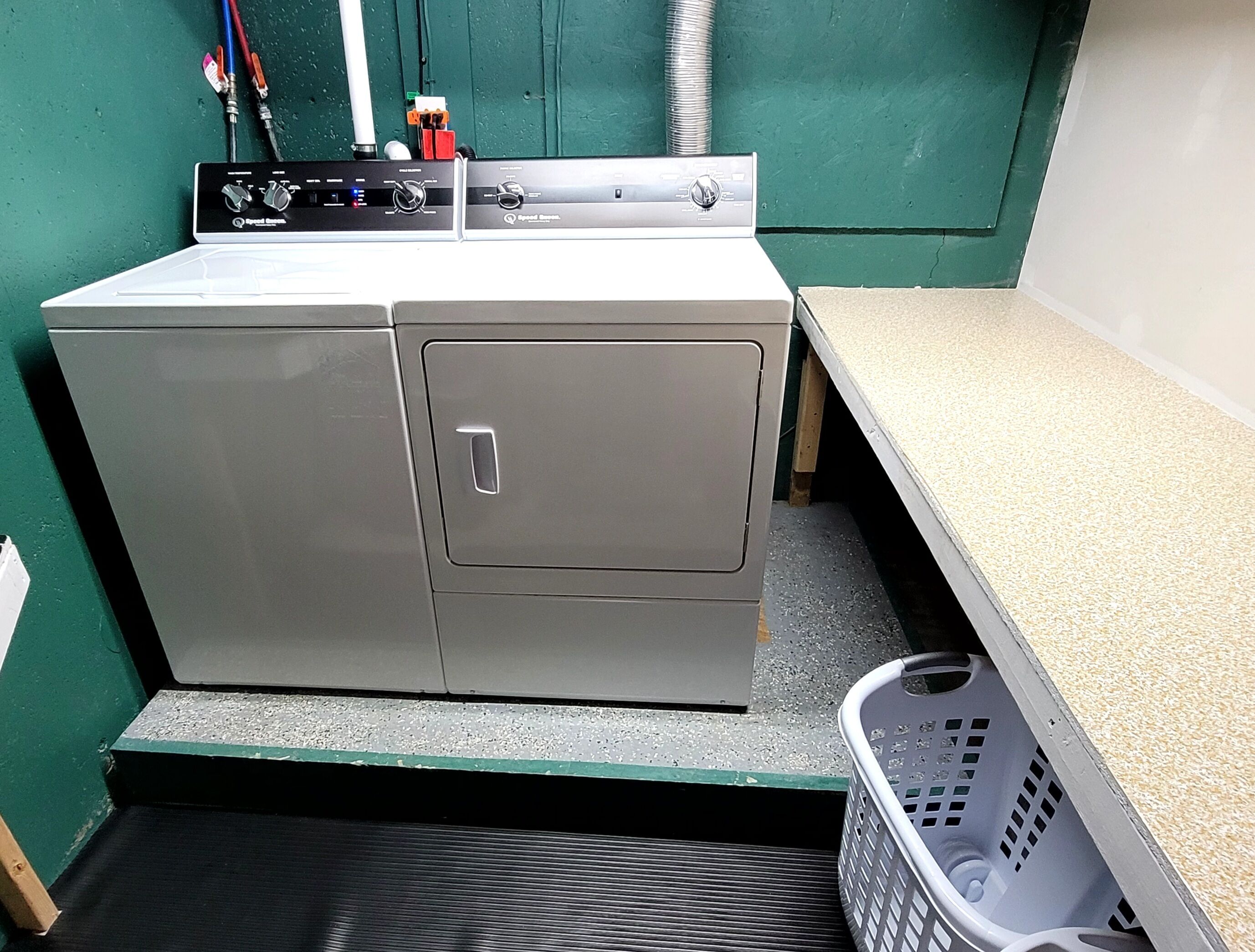 Lavatrice e asciugatrice a carica frontale, compatte e in acciaio inossidabile, con pannello di controllo, situate in una piccola lavanderia con parete verde e piano di lavoro color beige.