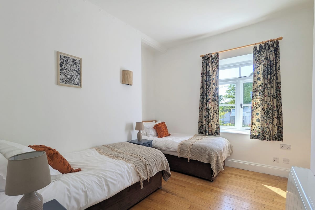 A bedroom with two beds, a window with floral curtains, a bedside lamp, and a small table with a lamp.