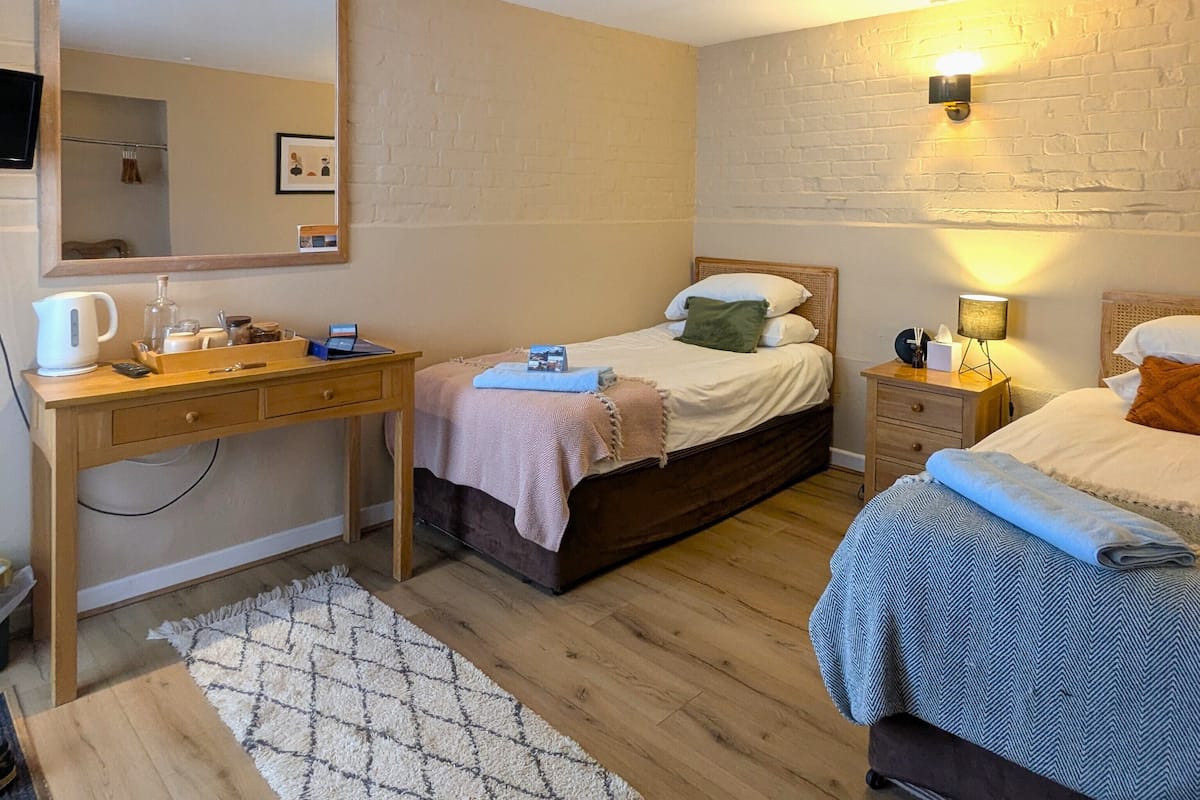 A bedroom with a wooden desk, a bed with a headboard, a bedside table with a lamp, and a wall-mounted light fixture.