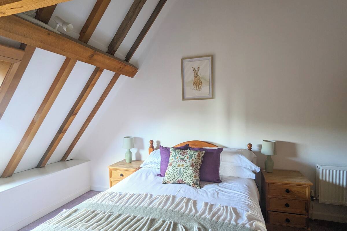 A bedroom with a sloped ceiling, a bed with white bedding and multiple pillows, two bedside tables with lamps, and a framed picture on the wall.