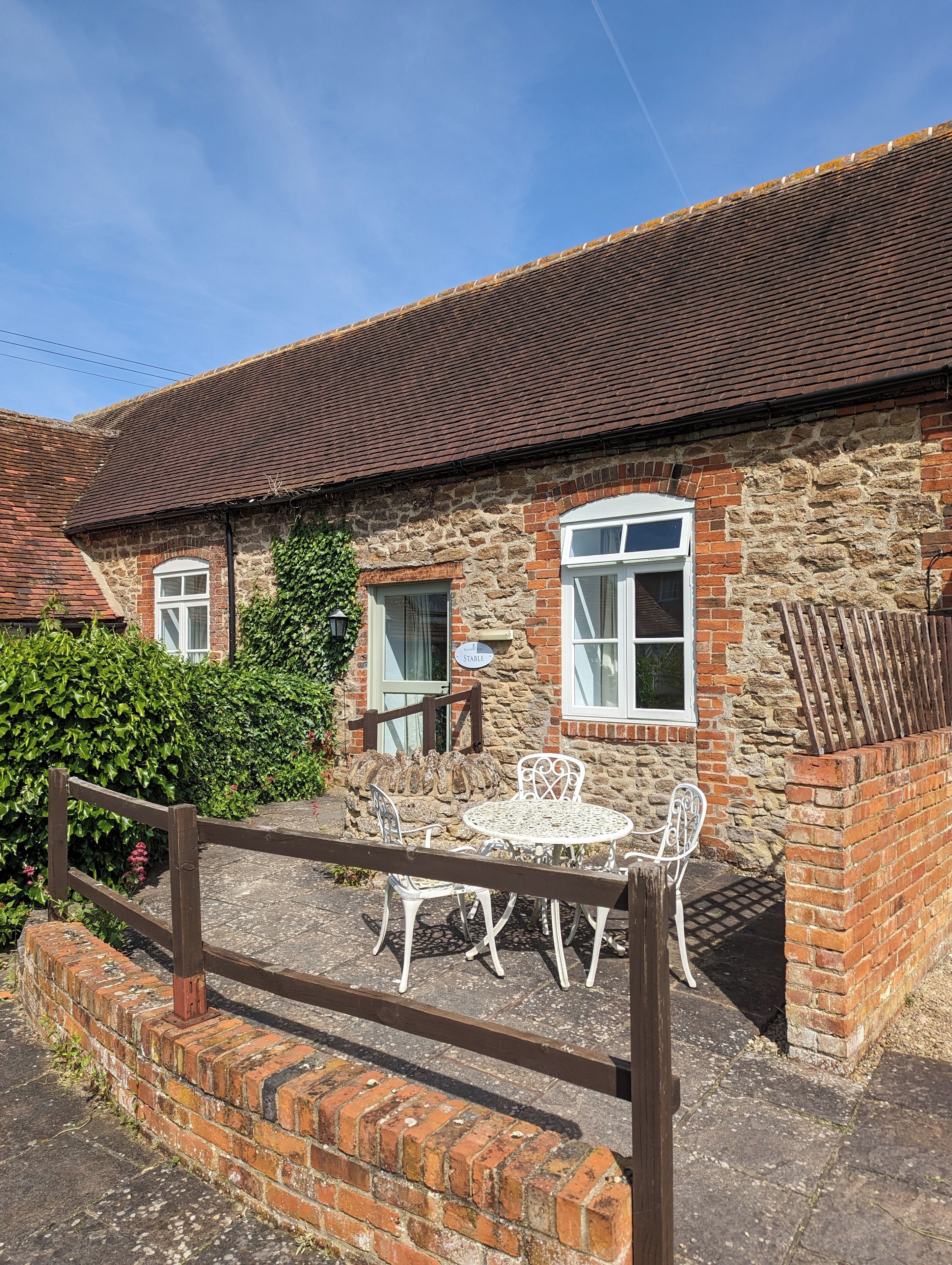 A stone building with a tiled roof, a brick entrance, and a small patio with white metal furniture.