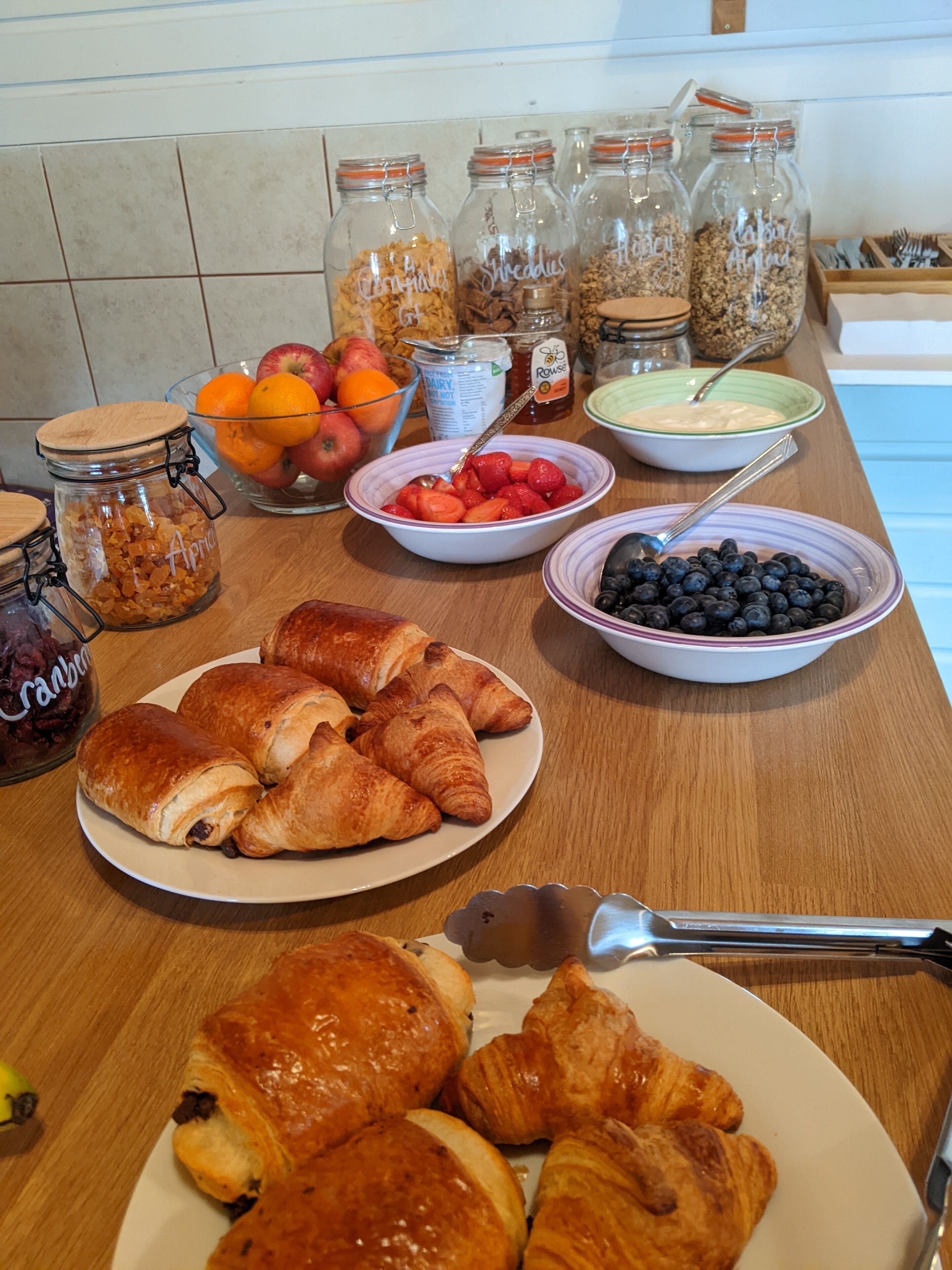 Una mesa de desayuno con pasteles, frutas y frascos de conservas.