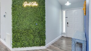 A hallway with a green wall covered in foliage, a neon sign that reads "Good Vibes," and a door with a blue accent wall.