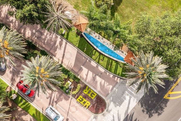 An aerial view of a landscaped area with a swimming pool, palm trees, and a road with marked parking spaces.