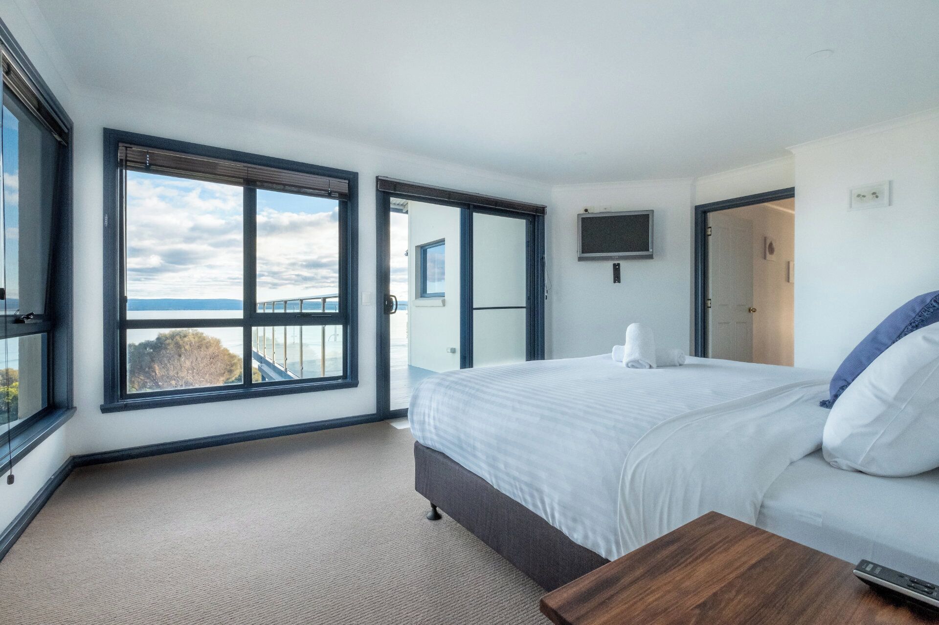 Ein modernes Hotelzimmer mit einem großen Bett, einem Holznachttisch, einem an der Wand befestigten Fernseher und einem Balkon mit Meerblick.
