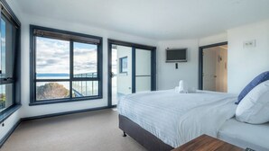 A modern hotel room with a large bed, a wooden side table, a TV mounted on the wall, and a balcony with a view of the sea.