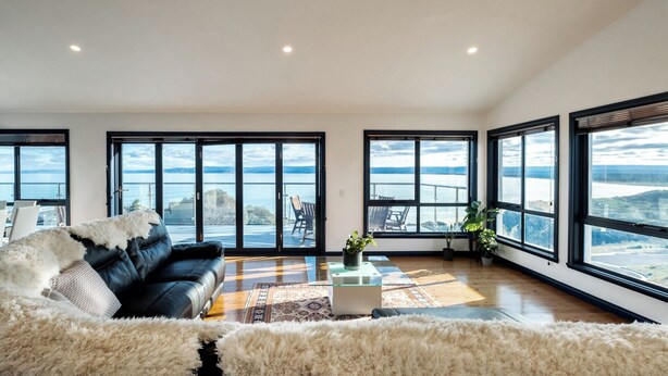 A spacious living room with a black leather sofa, a white furry rug, and a glass coffee table. Large windows offer a view of a lake and mountains.