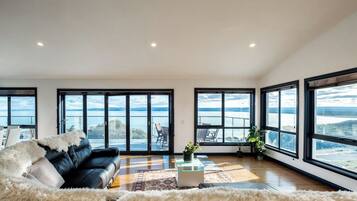 Ein geräumiges Wohnzimmer mit einem schwarzen Ledersofa, einem weißen, flauschigen Teppich und einem Glastisch. Große Fenster bieten Blick auf einen See und Berge.