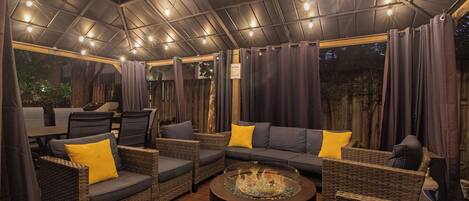 An outdoor seating area with wicker furniture and cushions, a glass-top coffee table, and a ceiling with string lights.
