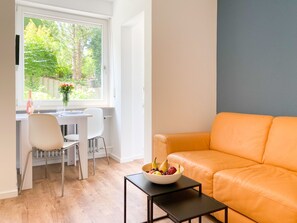 A modern living room with a leather sofa, a dining table with chairs, and a window with a view of greenery. - Cozy apartment with terrace in a quiet location (Kempten (Allgäu))