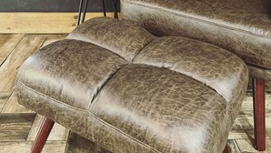 A leather sofa with matching ottoman, a small wooden side table with a glass vase containing a plant, and a textured wall in the background.