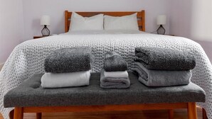 Cama bem arrumada com edredom branco, dois travesseiros e uma pilha de toalhas dobradas em um banco.