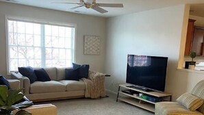 Un salon avec un canapé beige, un fauteuil à rayures, un téléviseur posé sur un meuble, un ventilateur de plafond et une fenêtre donnant sur des arbres.