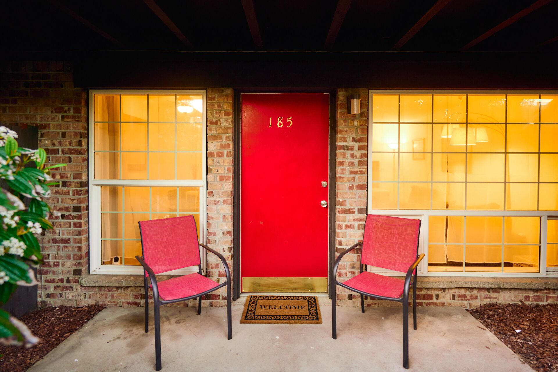 Een rode deur met het nummer 185, twee rode stoelen en een welkomstmat voor een gebouw met grote ramen.