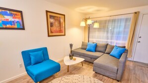 Ein Wohnzimmer mit einem blauen Sofa, einem grauen Sofa, einem kleinen weißen Tisch und einem gemusterten Teppich. An der Wand hängt ein Gemälde und ein Fenster ist mit Vorhängen versehen.