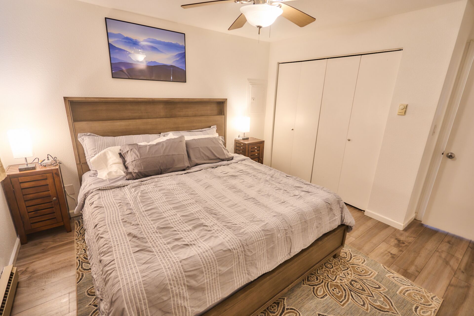 A bedroom with a large bed, a ceiling fan, a nightstand with a lamp, a wall-mounted TV, and a wardrobe.
