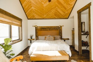 A bedroom with a wooden ceiling, a large bed with a fluffy white comforter, a wooden bench, and a large window with blinds. - Luxury ranch cabin for 12, dog friendly with hot tub, private valley with creek! (Creede)