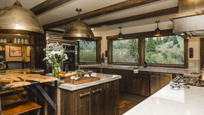 Une cuisine avec des poutres en bois, de grandes suspensions, un îlot central doté d’une planche à découper et une vue sur la verdure à l’extérieur.