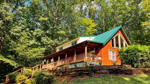 A log cabin with a green roof, a porch, and a staircase leading up to it, surrounded by dense trees.