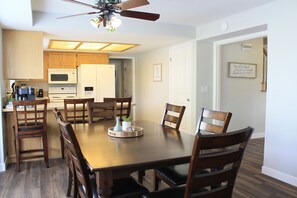 A dining area with a wooden table and chairs, a ceiling fan, and a kitchen in the background.